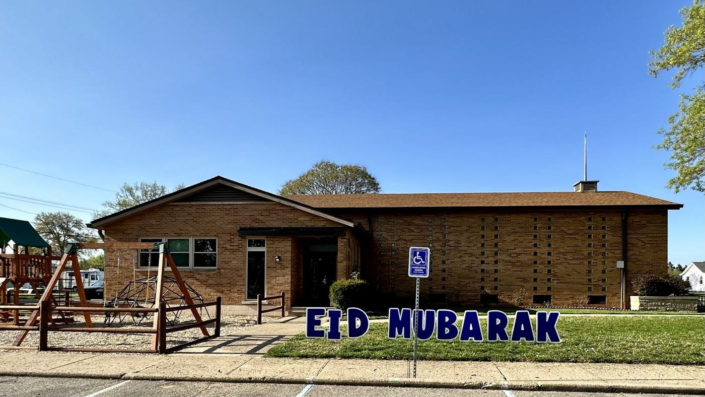 The brick masjid on Chestnut Street seen from the parking lot, with Eid Mubarak letters on the lawn and the play set to the left